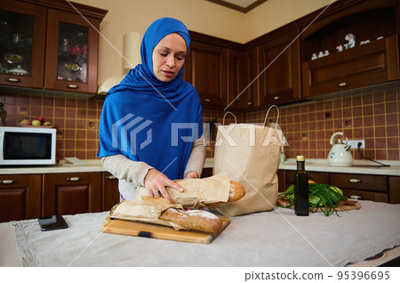 Muslim Middle Eastern pretty woman housewife in blue hijab, puts whole grain sourdough bread loaves on a wooden board, while unpacking fresh food from paper grocery bag in the home kitchen Muslim Middle Eastern pretty woman housewife in blue hijab, puts whole grain sourdough bread loaves on a wooden board, while unpacking fresh food from paper grocery bag in the home kitchen 95396695