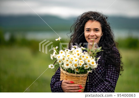 A woman stands on a green field and holds a basket with a large bouquet of daisies in her hands. In the background are mountains and a lake. A woman stands on a green field and holds a basket with a large bouquet of daisies in her hands. In the background are mountains and a lake. 95400863