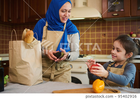 Caucasian little girl helps her mother unpack the delivered vegetables, takes out bell pepper, fresh clean paprika from paper bag. Muslim woman and her daughter arrive home after grocery shopping Caucasian little girl helps her mother unpack the delivered vegetables, takes out bell pepper, fresh clean paprika from paper bag. Muslim woman and her daughter arrive home after grocery shopping 95402916