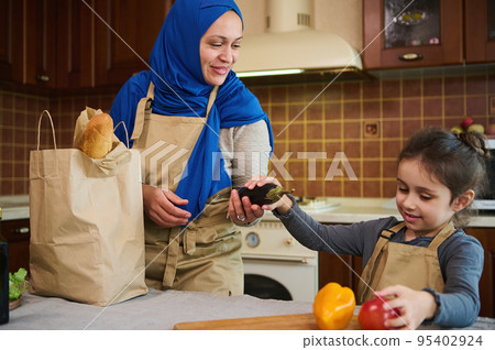 Cheerful happy Middle Eastern Muslim woman housewife in blue hijab, a loving mother and her lovely daughter unpacking grocery bag after shopping. Customers of online supermarkets. Food delivery Cheerful happy Middle Eastern Muslim woman housewife in blue hijab, a loving mother and her lovely daughter unpacking grocery bag after shopping. Customers of online supermarkets. Food delivery 95402924