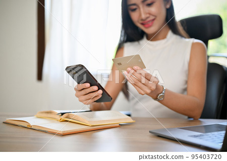 Attractive Asian businesswoman at her desk, holding a mobile phone and a credit card Attractive Asian businesswoman at her desk, holding a mobile phone and a credit card 95403720