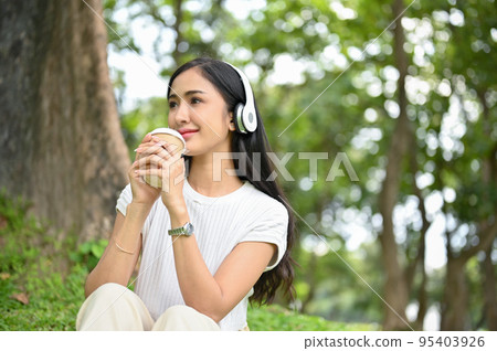 Attractive Asian female listening to music and sipping coffee while relaxing in park. Attractive Asian female listening to music and sipping coffee while relaxing in park. 95403926