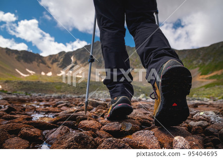 Hiking boots close-up on mountain rocks by the lake with trekking poles, view from below Hiking boots close-up on mountain rocks by the lake with trekking poles, view from below 95404695
