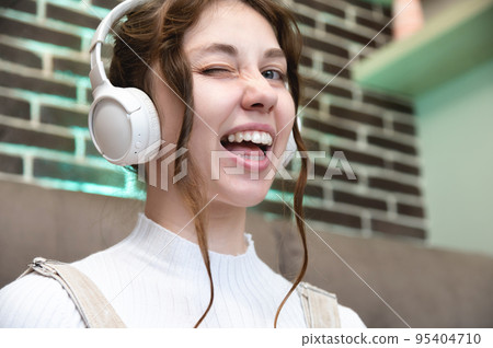 Young caucasian teenage woman smiles and winks, she listens to music in headphones while sitting at home Young caucasian teenage woman smiles and winks, she listens to music in headphones while sitting at home 95404710