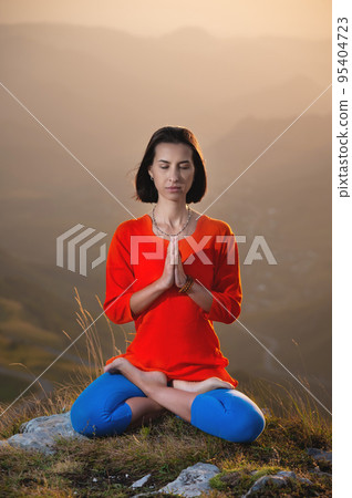 young caucasian woman meditates in the lotus position in the grass in the mountains near the cliff in the setting sun 95404723