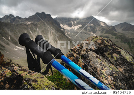 trekking poles close-up, lie on a stone overgrown with grass, in summer, against the backdrop of snow-capped rocky mountains 95404739