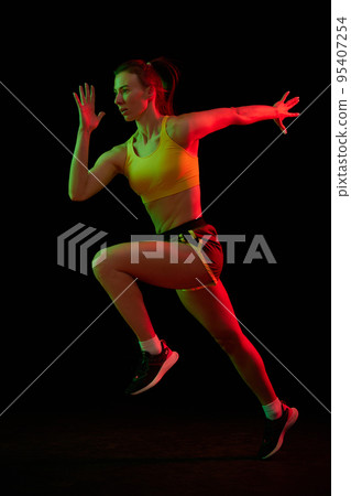 Dynamic portrait of young sportive woman training, running isolated on dark background in neon light. Competition, sport, fitness, speed and energy concept. Dynamic portrait of young sportive woman training, running isolated on dark background in neon light. Competition, sport, fitness, speed and energy concept. 95407254