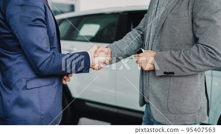 Male worker of car showroom is giving car keys to buyer stylish young man and shaking hands with him standing beside luxurious new car. Selling and buying vehicles concept. Male worker of car showroom is giving car keys to buyer stylish young man and shaking hands with him standing beside luxurious new car. Selling and buying vehicles concept. 95407563