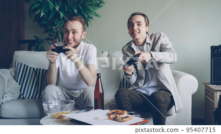 Two handsome young guys are playing videogame holding joysticks sitting on sofa at home. Emotional men are enjoying game, snacks and beer bottles are visible. Two handsome young guys are playing videogame holding joysticks sitting on sofa at home. Emotional men are enjoying game, snacks and beer bottles are visible. 95407715