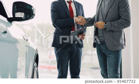 Motor showroom employee is giving car key to young male buyer and shaking hands, dealer is holding documents and gesturing. Selling and buying luxury transport concept. 95407732