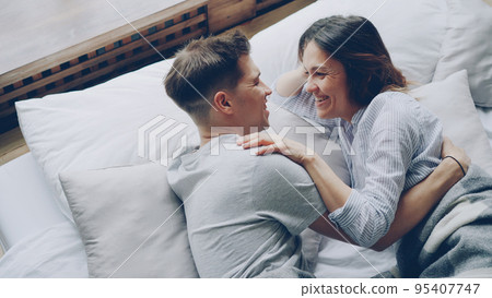 Attractive asian woman is lying in bed with her boyfriend touching him while he is hugging her, they are talking and laughing. Tenderness and love concept. 95407747