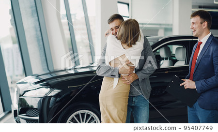 Young bearded man is buying car for his attractive wife, she is kissing and hugging him. 95407749