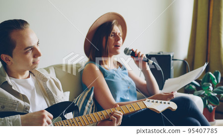 Creative musical duet is practising at home attractive woman is singing in microphone and handsome man is playing the guitar. Young cheerful people are wearing casual clothes. 95407785