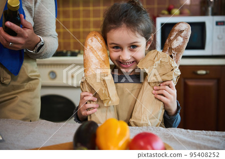 Adorable Caucasian child, funny little girl with two loaves of whole grain sourdough bread, smiling with a cheerful toothy smile, looking at camera, standing at kitchen table, unpacking grocery bag Adorable Caucasian child, funny little girl with two loaves of whole grain sourdough bread, smiling with a cheerful toothy smile, looking at camera, standing at kitchen table, unpacking grocery bag 95408252