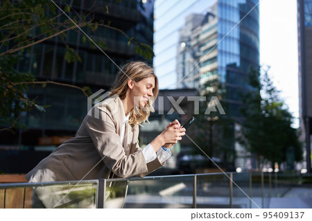 Smiling businesswoman using her mobile phone on a break, outside of office, looking at smartphone screen while messaging someone 95409137