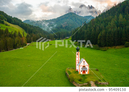 Aerial view of St. Johann chapel in South Tyrol against the Dolomites covered by clouds at sunset.  95409138