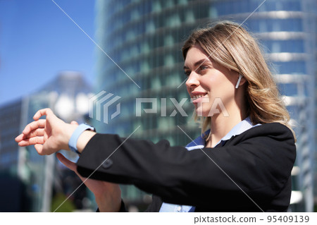 Portrait of busy corporate woman in wireless headphones, looking at time on her digital watch, checking messages, adjusting music, standing outdoors on street 95409139