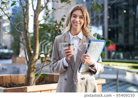 Young businesswoman standing on street with takeaway coffee, laptop and work documents, smiling Young businesswoman standing on street with takeaway coffee, laptop and work documents, smiling 95409150