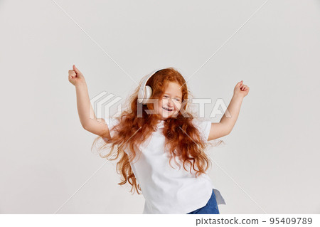 Joyful little girl, kid in white t-shirt and jeans listening to music and dancing isolated over white background. Kids fashion, emotions, carefree childhood 95409789