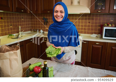 Beautiful Arab Muslim woman, pleasant housewife with her head covered in a blue hijab, holding fresh salad leaves while preparing raw vegan healthy meal, in the home kitchen. Cooking. Vegetarianism. Beautiful Arab Muslim woman, pleasant housewife with her head covered in a blue hijab, holding fresh salad leaves while preparing raw vegan healthy meal, in the home kitchen. Cooking. Vegetarianism. 95409929