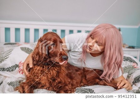 Beautiful girl lying relax in bed with Irish Setter dog. Morning prodding in a warm comfortable bed.Cute woman and her dog are sleeping in the bed covered with a blanket Beautiful girl lying relax in bed with Irish Setter dog. Morning prodding in a warm comfortable bed.Cute woman and her dog are sleeping in the bed covered with a blanket 95412351