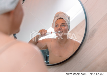 Young caucasian woman brushing teeth with electric toothbrush in bathroom. Modern domestic lifestyle. Hygiene and dental care Young caucasian woman brushing teeth with electric toothbrush in bathroom. Modern domestic lifestyle. Hygiene and dental care 95412365