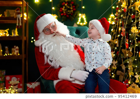 Funny boy checking the red nose of Santa Claus while sitting on his lap. New Year concept  95412469