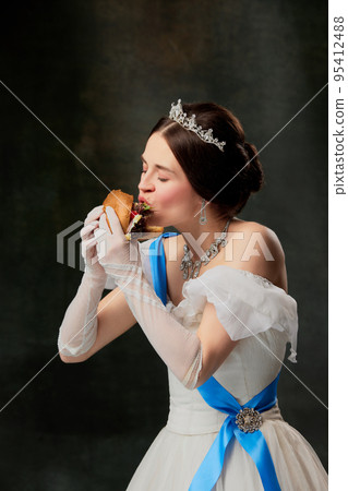 Enjoy the taste. Beautiful young queen or princess in white medieval outfit eating burger on dark vintage background. Art, creativity, beauty, taste Enjoy the taste. Beautiful young queen or princess in white medieval outfit eating burger on dark vintage background. Art, creativity, beauty, taste 95412488