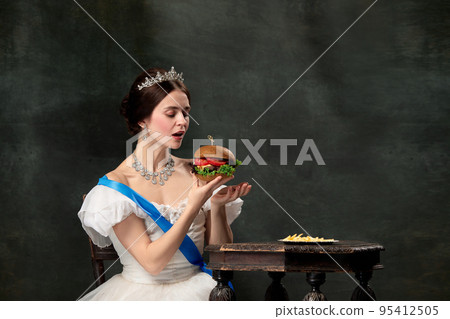 Wow. Young beautiful woman, royal person, queen or princess in white medieval outfit tasting burger on dark background. Comparison of eras, modern, fashion, beauty. Wow. Young beautiful woman, royal person, queen or princess in white medieval outfit tasting burger on dark background. Comparison of eras, modern, fashion, beauty. 95412505