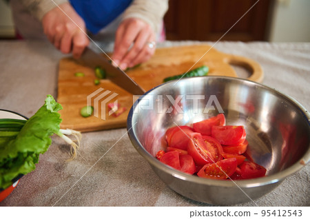 Selective focus on a metal bowl with chopped ripe juicy red tomatoes on a kitchen table with linen tablecloth, against a blurred background of woman's hands slicing cucumbers on a wooden cutting board 95412543