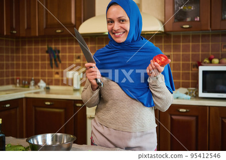 Charming Arabic Muslim woman housewife in blue hijab, smiling a toothy smile, looking at camera, standing with a kitchen knife and ripe fresh juicy tomato in her hands, against home kitchen background 95412546