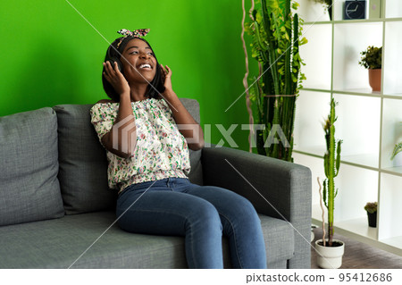 African woman listening to music with headphones while sitting on couch at home African woman listening to music with headphones while sitting on couch at home 95412686