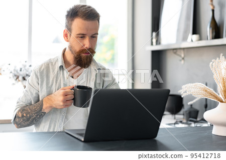Stylish bearded man working on laptop from home sitting at the table 95412718