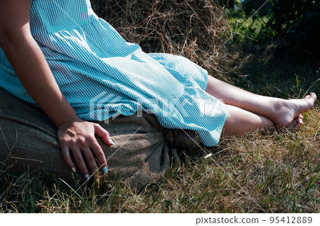 A barefoot girl in a blue cotton dress sits on a bag of hay in nature. Summer relax on the hay. Clothed at the farm. A barefoot girl in a blue cotton dress sits on a bag of hay in nature. Summer relax on the hay. Clothed at the farm. 95412889