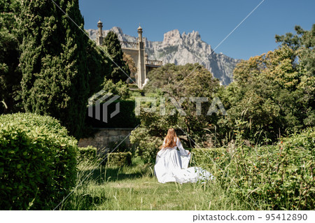 a beautiful woman with long brown hair and long white dress stay along a path along beautiful bushes in the park rear view a beautiful woman with long brown hair and long white dress stay along a path along beautiful bushes in the park rear view 95412890