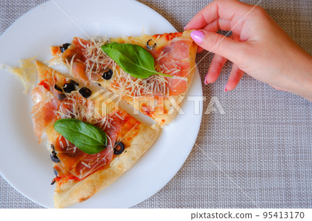 Close-up top view of a female hand takes a slice of pizza from a white plate. Appetizing delicious pizza. 95413170