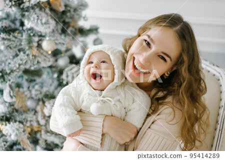 Portrait of a happy mother and a charming child celebrating Christmas. Smiling baby with mom in a festively decorated room with a Christmas tree and decorations. 95414289