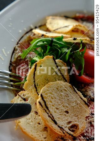 Beef Carpaccio cold appetizer with parmesan, tomatos and arugula on white plate with hardtack on dark background Beef Carpaccio cold appetizer with parmesan, tomatos and arugula on white plate with hardtack on dark background 95415429