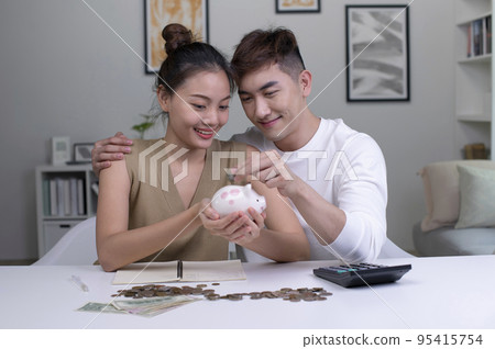 Asian couple at home saving money in a piggybank and smiling. 95415754