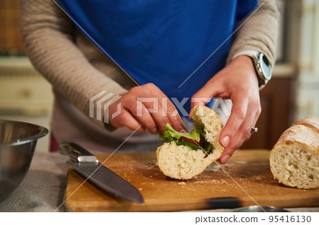 Details: Woman's hands put greens and organic fresh vegetables in the bread, making crusty sandwiches with veggies in the home kitchen, preparing a delicious healthy snack 95416130