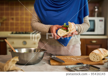 Details: Hands of housewife, with head covered in blue hijab, holding loaves of whole grain sourdough bread and making sandwiches with veggies in the home kitchen, preparing a delicious healthy snack Details: Hands of housewife, with head covered in blue hijab, holding loaves of whole grain sourdough bread and making sandwiches with veggies in the home kitchen, preparing a delicious healthy snack 95416143