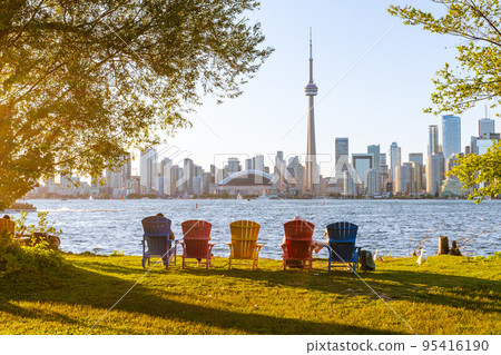 Colorful adirondack chairs on Toronto Island Park at sunset time. Toronto City downtown skyline in the background. Ontario, Canada. Colorful adirondack chairs on Toronto Island Park at sunset time. Toronto City downtown skyline in the background. Ontario, Canada. 95416190