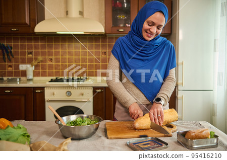 Pleasant Middle-Eastern Muslim woman, housewife in a blue hijab cutting loaf of whole grain sourdough wheat bread on a wooden board, making sandwiches in a cozy home kitchen with rustic wooden design 95416210