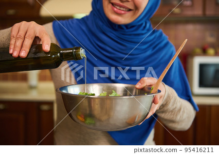 Close-up of a blurred Muslim woman in hijab, smiling a beautiful toothy smile, pouring olive oil into a bowl with vegetable salad. Cookery concept. Selective focus housewife cooking healthy dinner 95416211