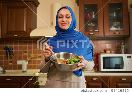 Charming Muslim Arab young woman with head covered in blue hijab, standing in the home kitchen with a bowl of healthy vegetable salad for dinner, smiling a beautiful toothy smile looking at the camera 95416215