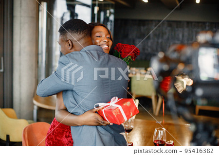 Black man gifted a bouquet of red roses and gift box for his girlfriend. Woman wearing red elegant dress and man blue costume. Couple hugging while standing at restaurant. Black man gifted a bouquet of red roses and gift box for his girlfriend. Woman wearing red elegant dress and man blue costume. Couple hugging while standing at restaurant. 95416382
