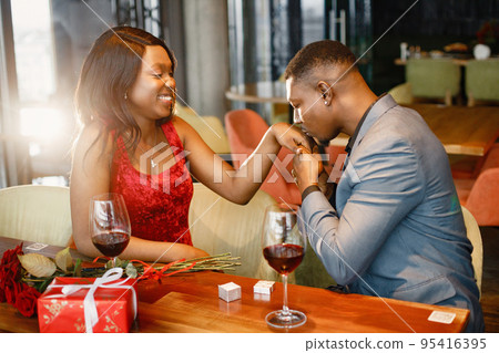 Couple enjoying day out at restaurant. Black man kissing a hand of his black girlfriend. Woman wearing red elegant dress and man blue costume. Couple enjoying day out at restaurant. Black man kissing a hand of his black girlfriend. Woman wearing red elegant dress and man blue costume. 95416395