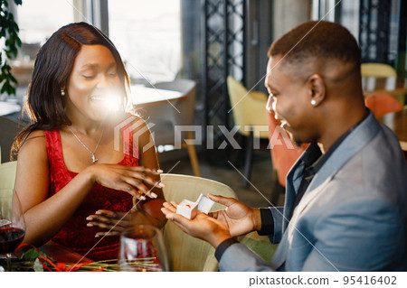 Couple enjoying day out at restaurant. Black man holding a little box with engagement ring. Woman wearing red elegant dress and man blue costume. 95416402
