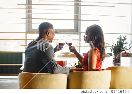 Backside photo of a couple enjoying day out at restaurant. Black man and woman drinking wine. Woman wearing red elegant dress and man blue costume. 95416404
