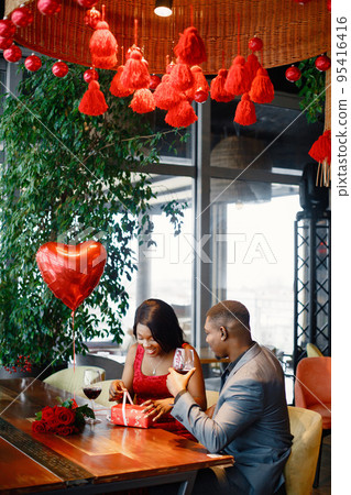 Couple enjoying day out at restaurant. Black man and woman drinking wine. Woman wearing red elegant dress and opening a gift box near a boiquet of red roses. 95416416